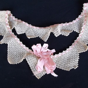 May include: Cream-colored lace collar with a scalloped edge, accented with a delicate pink ribbon. A matching pink bow is centered on the collar. The collar is displayed against a dark background.