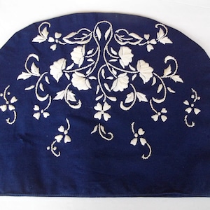 May include: A navy blue teapot cozy with white embroidered floral designs.