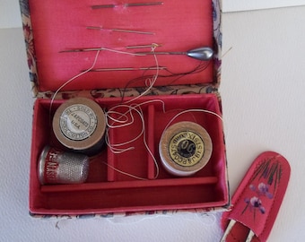 May include: Vintage sewing kit with a red velvet interior and a floral patterned exterior. The kit contains needles, spools of thread, a thimble, and a small scissor holder. The kit is open, revealing the contents, and is set against a white background.