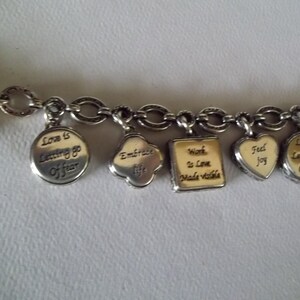 K&ouml;nnte beinhalten: Silbernes Charm-Armband mit verschiedenen Anh&auml;ngern. Zu den Anh&auml;ngern geh&ouml;ren ein runder Anh&auml;nger mit dem Text "Love is Letting go of fear", ein Kleeblatt-Anh&auml;nger mit "Embrace life" und ein Herz-Anh&auml;nger mit "Feel joy".