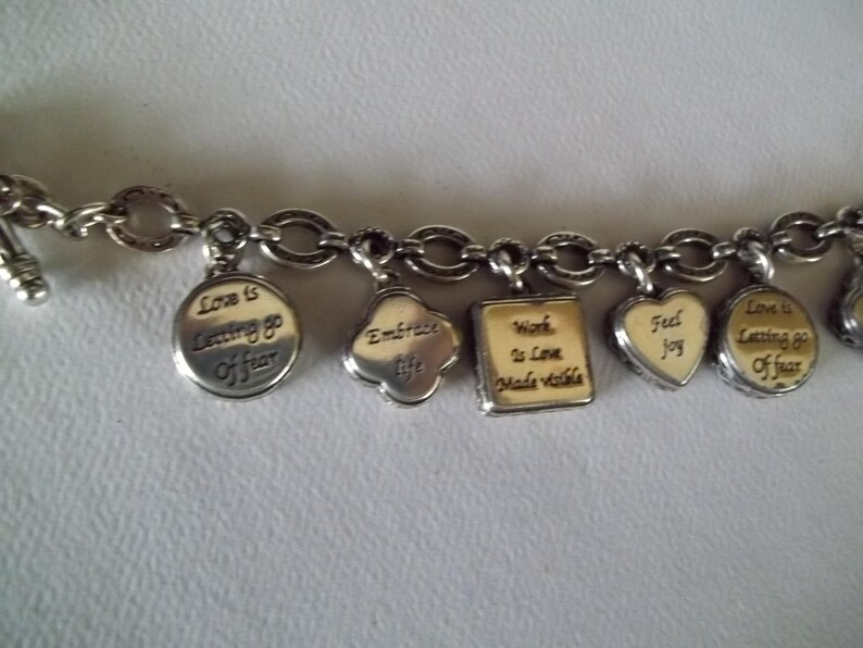 K&ouml;nnte beinhalten: Silbernes Charm-Armband mit verschiedenen Anh&auml;ngern. Zu den Anh&auml;ngern geh&ouml;ren ein runder Anh&auml;nger mit dem Text "Love is Letting go of fear", ein Kleeblatt-Anh&auml;nger mit "Embrace life" und ein Herz-Anh&auml;nger mit "Feel joy".