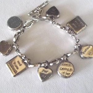 K&ouml;nnte beinhalten: Silbernes Charm-Armband mit verschiedenen Anh&auml;ngern in verschiedenen Formen, darunter Herzen, Quadrate und Kreise. Einige Anh&auml;nger haben goldfarbene Akzente und inspirierende S&auml;tze wie "Embrace Life" und "Love is Letting Go of Fear".