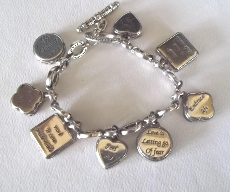 K&ouml;nnte beinhalten: Silbernes Charm-Armband mit verschiedenen Anh&auml;ngern in verschiedenen Formen, darunter Herzen, Quadrate und Kreise. Einige Anh&auml;nger haben goldfarbene Akzente und inspirierende S&auml;tze wie "Embrace Life" und "Love is Letting Go of Fear".