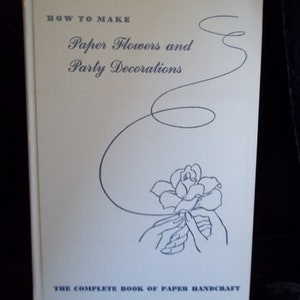 以下が含まれることがあります： 「How to Make Paper Flowers and Party Decorations: The Complete Book of Paper Handcraft」というタイトルの書籍。青い紙の花を持った手のイラストが描かれている。