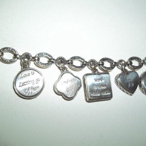 K&ouml;nnte beinhalten: Silbernes Charm-Armband mit verschiedenen Anh&auml;ngern. Zu den Anh&auml;ngern geh&ouml;ren ein runder Anh&auml;nger mit dem Text "Love is Letting go Of fear", ein Kleeblatt-Anh&auml;nger, ein quadratischer Anh&auml;nger mit dem Text "Work I Love Make a Difference" und herzf&ouml;rmige Anh&auml;nger.