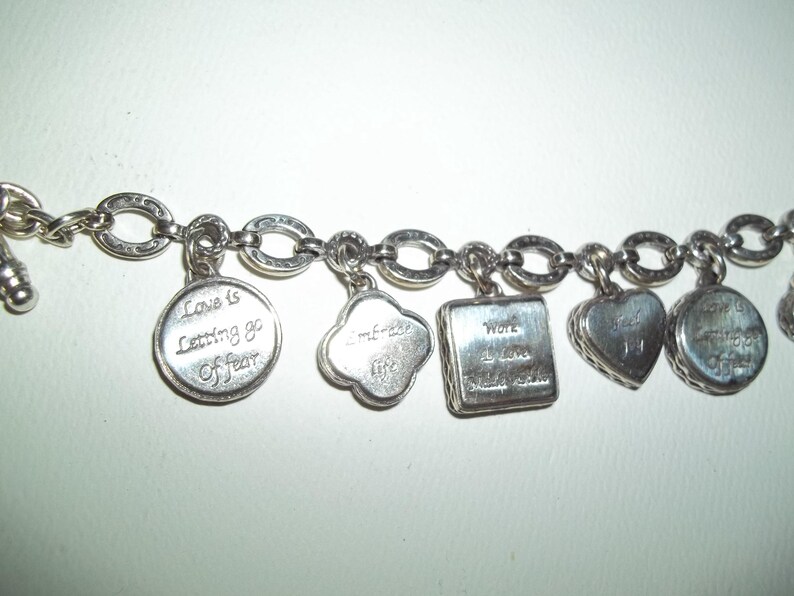 K&ouml;nnte beinhalten: Silbernes Charm-Armband mit verschiedenen Anh&auml;ngern. Zu den Anh&auml;ngern geh&ouml;ren ein runder Anh&auml;nger mit dem Text "Love is Letting go Of fear", ein Kleeblatt-Anh&auml;nger, ein quadratischer Anh&auml;nger mit dem Text "Work I Love Make a Difference" und herzf&ouml;rmige Anh&auml;nger.