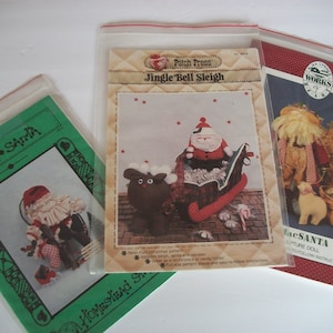 Puede incluir: Tres patrones de artesanía con temática navideña en bolsas de plástico. Los patrones son para un muñeco de Papá Noel, un trineo con cascabeles y un muñeco de Papá Noel. Los patrones son de Patch Press y Small Works of Art.