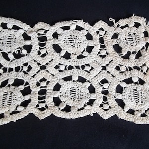 May include: Cream-colored, handmade lace doily with a repeating floral pattern. The lace features circular and oval shapes connected by delicate openwork. The edges are scalloped, adding a decorative touch. The doily is likely made of cotton or a similar fiber.