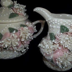 Könnte beinhalten: Ein weißes Porzellan-Zucker- und Milchkännchen-Set mit rosa und weißen floralen Akzenten. Das Set ist mit weißen Punkten und grünen Blättern verziert.