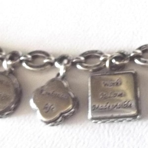 K&ouml;nnte beinhalten: Silbernes Charm-Armband mit verschiedenen Anh&auml;ngern. Zu den Anh&auml;ngern geh&ouml;ren ein Herz, ein Buch, ein Kleeblatt und ein Quadrat mit den Worten "Feel Joy" und anderen inspirierenden S&auml;tzen. Das Armband hat einen ovalen Verschluss.