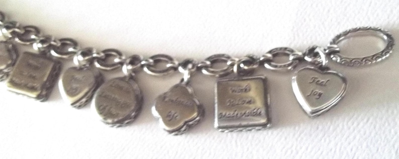 K&ouml;nnte beinhalten: Silbernes Charm-Armband mit verschiedenen Anh&auml;ngern. Zu den Anh&auml;ngern geh&ouml;ren ein Herz, ein Buch, ein Kleeblatt und ein Quadrat mit den Worten "Feel Joy" und anderen inspirierenden S&auml;tzen. Das Armband hat einen ovalen Verschluss.