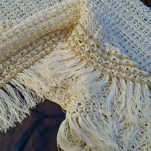 May include: A cream and tan crocheted shawl featuring an openwork lattice pattern. The textile has a decorative border with long, loose cream-colored fringe tassels along the edge, creating a textured and layered appearance across the fabric.