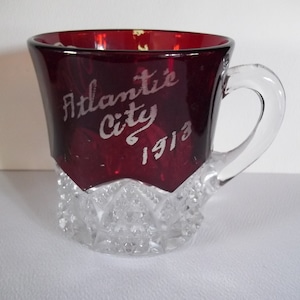 Puede incluir: Una taza de recuerdo de vidrio rojo con una base de vidrio transparente y tallada. La taza tiene un asa y dice "Atlantic City 1913" en letras blancas.