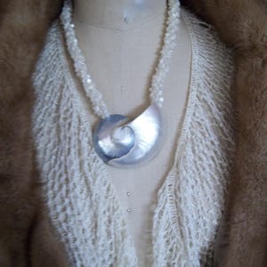 May include: A white crocheted shawl with a white beaded necklace and a large silver nautilus shell pendant.
