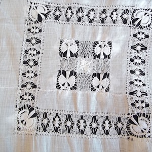 Op de afbeelding: Close-up van een wit, vierkant, geborduurd textiel met ingewikkelde kanten details. Het ontwerp heeft bloemen- en geometrische patronen, met een centraal vierkant en randen van delicate kant. De achtergrond is zwart, wat de witte borduursels benadrukt.