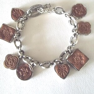 K&ouml;nnte beinhalten: Silberfarbenes Charm-Armband mit verschiedenen kupferfarbenen Anh&auml;ngern. Die Anh&auml;nger umfassen Quadrate, Kreise, Herzen und Blumenformen. Das Armband hat einen Karabinerverschluss und liegt auf einer wei&szlig;en Oberfl&auml;che.