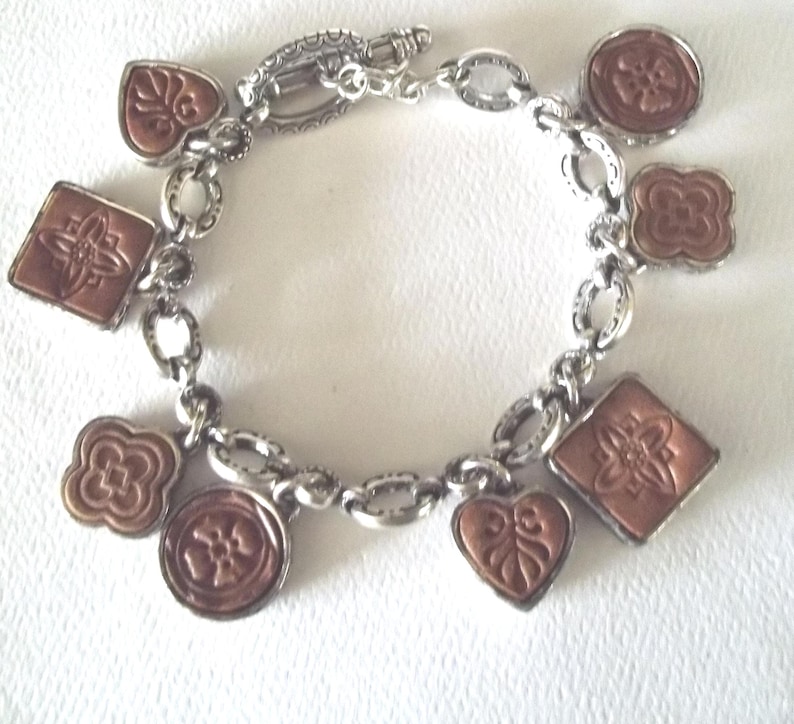 K&ouml;nnte beinhalten: Silberfarbenes Charm-Armband mit verschiedenen kupferfarbenen Anh&auml;ngern. Die Anh&auml;nger umfassen Quadrate, Kreise, Herzen und Blumenformen. Das Armband hat einen Karabinerverschluss und liegt auf einer wei&szlig;en Oberfl&auml;che.