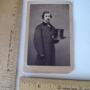 May include: A vintage black and white photograph of a man in a long coat, holding a top hat. The man has a mustache and is wearing a dark suit with a bow tie. The photograph is rectangular and has a sepia tone.