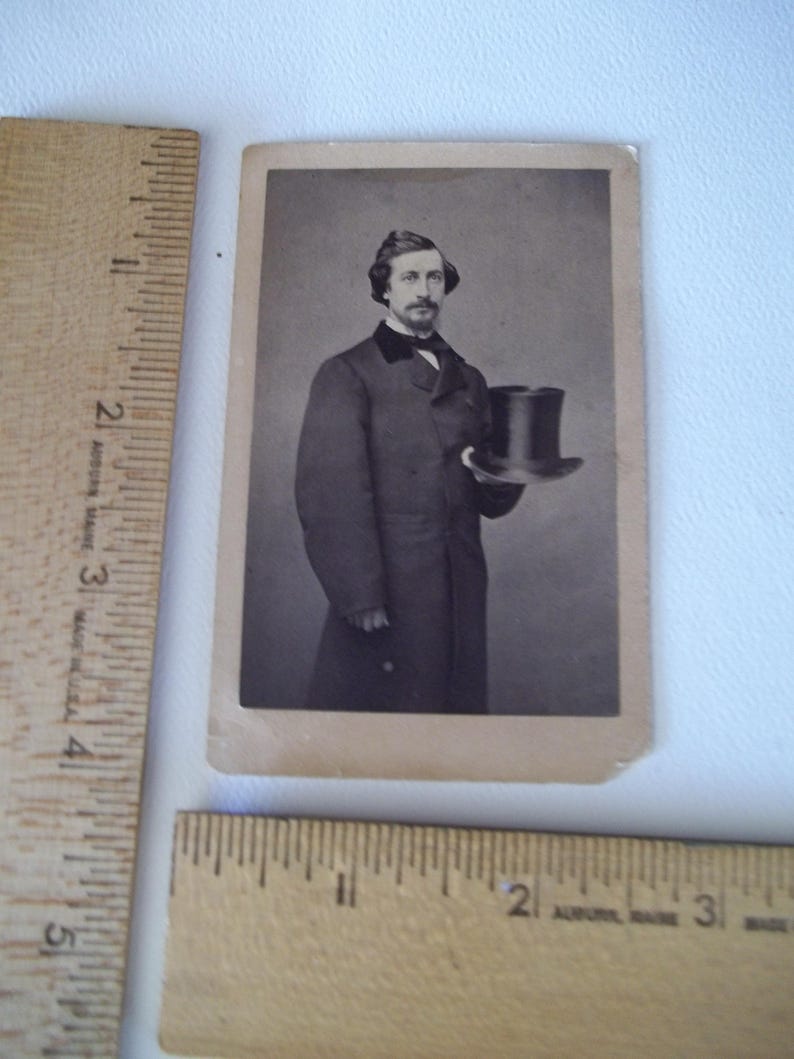 May include: A vintage black and white photograph of a man in a long coat, holding a top hat. The man has a mustache and is wearing a dark suit with a bow tie. The photograph is rectangular and has a sepia tone.