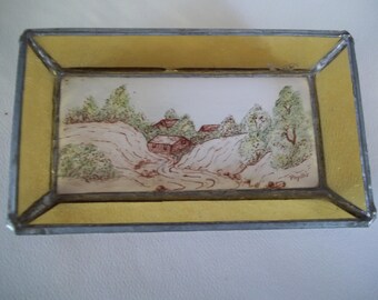 Decoración de pared vintage con escena de pueblo hecha a mano en azulejos pequeños y laterales de vidrieras en tonos beige y amarillo.