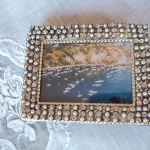 May include: A rectangular picture frame featuring a metallic border encrusted with alternating clear rhinestones and small white pearls. The frame contains a landscape painting depicting a coastal scene with cliffs and boats on blue water.
