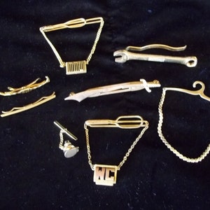 May include: A collection of gold-toned tie clips and tie bars. The tie clips feature a variety of designs, including a wrench, a sword, a pair of skis, and a hanger. The tie bars feature a chain with a rectangular charm that says "WC".