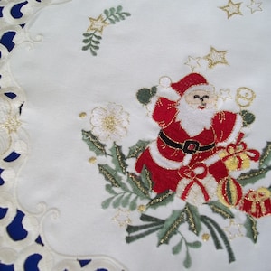 Può includere: Tovaglia da tavolo natalizia bianca ricamata con un Babbo Natale rosso e bianco che tiene un sacchetto regalo. Il design include agrifoglio, bacche e stelle.