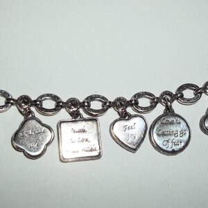 K&ouml;nnte beinhalten: Silbernes Charm-Armband mit verschiedenen Anh&auml;ngern in unterschiedlichen Formen, darunter Herzen und Kreise. Jeder Anh&auml;nger ist mit einer inspirierenden Botschaft graviert. Das Armband hat einen Knebelverschluss.