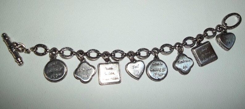 K&ouml;nnte beinhalten: Silbernes Charm-Armband mit verschiedenen Anh&auml;ngern in unterschiedlichen Formen, darunter Herzen und Kreise. Jeder Anh&auml;nger ist mit einer inspirierenden Botschaft graviert. Das Armband hat einen Knebelverschluss.