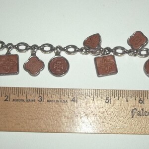 K&ouml;nnte beinhalten: Ein silberfarbenes Charm-Armband mit sieben braunen Anh&auml;ngern in verschiedenen Formen, darunter Herzen, Quadrate und Kreise. Das Armband ist etwa 18 cm lang, gemessen mit einem Holzlineal.
