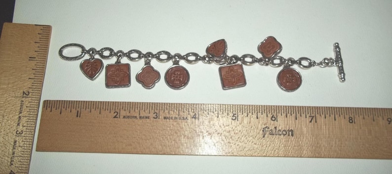 K&ouml;nnte beinhalten: Ein silberfarbenes Charm-Armband mit sieben braunen Anh&auml;ngern in verschiedenen Formen, darunter Herzen, Quadrate und Kreise. Das Armband ist etwa 18 cm lang, gemessen mit einem Holzlineal.