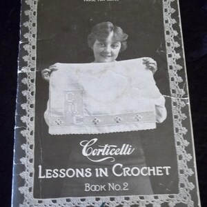 May include: A vintage black and white instructional booklet titled Corticelli Lessons in Crochet Book No. 2. The cover features a person holding a white crocheted piece with the letters CMC visible, framed by a decorative crocheted border pattern.