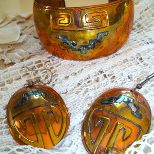 Op de afbeelding: Een koperkleurige manchetarmband en bijpassende oorbellen. De sieraden hebben een geometrisch ontwerp met gouden en oranje accenten en iriserende inlays. De manchet is breed en open, terwijl de oorbellen ovaal zijn met een haaksluiting.