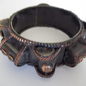 May include: A dark brown hinged bangle bracelet with copper-coloured accents. The bracelet features a series of rectangular and circular decorative elements, with a rope-like texture around the edges. Set against a white background.