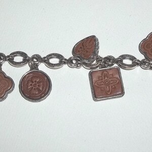 K&ouml;nnte beinhalten: Silberfarbenes Charm-Armband mit einer Reihe von Anh&auml;ngern in verschiedenen Formen, darunter Herzen, Quadrate und Kreise. Die Anh&auml;nger haben eine rotbraune Mitte mit detaillierten Designs. Das Armband hat einen Knebelverschluss.