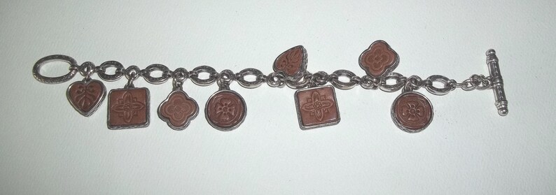 K&ouml;nnte beinhalten: Silberfarbenes Charm-Armband mit einer Reihe von Anh&auml;ngern in verschiedenen Formen, darunter Herzen, Quadrate und Kreise. Die Anh&auml;nger haben eine rotbraune Mitte mit detaillierten Designs. Das Armband hat einen Knebelverschluss.