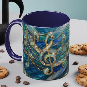 May include: A ceramic coffee mug with a navy blue interior and handle. The exterior features a stained-glass design with musical notes in gold and shades of blue and green. Chocolate chip cookies and coffee beans are scattered around the mug.