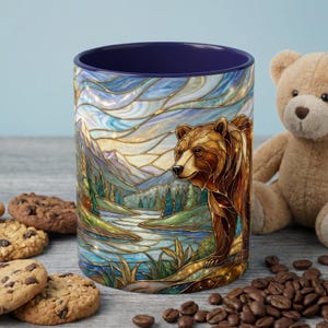 May include: A ceramic mug with a stained-glass style design featuring a brown bear in a mountain landscape. The mug has a dark blue interior and is surrounded by cookies, coffee beans, and a teddy bear.