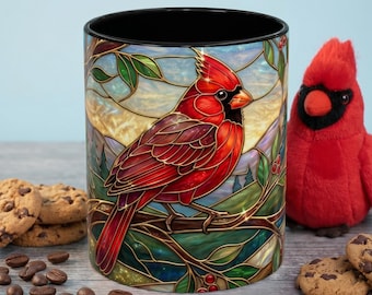 May include: A ceramic mug with a stained-glass design featuring a red cardinal bird perched on a branch. The mug has a black interior and is surrounded by cookies, coffee beans, and a plush cardinal toy.