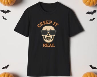 May include: Black t-shirt with a skull wearing sunglasses graphic and the text "CREEP IT REAL" in a Halloween theme. The design is in tan and black. The shirt is surrounded by pumpkins and bat decorations.