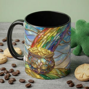 Puede incluir: Una taza de café con asa negra con un diseño de vidriera que presenta un arcoíris, una olla de monedas de oro y un cielo azul. La taza está rodeada de granos de café y galletas.