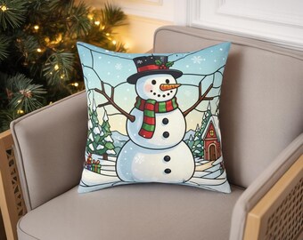 Op de afbeelding: Een decoratief kussen met een sneeuwmanontwerp. De sneeuwman heeft een zwarte hoge hoed, een wortelneus en een rood-groene sjaal. De achtergrond toont een winterlandschap met sneeuw, bomen en een klein huis.