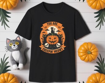 Puede incluir: Camiseta negra con un diseño de Halloween. El diseño presenta una calabaza tallada, gatos negros y el texto "YOU ARE CREEPIN' MEOWT". La camiseta está rodeada de calabazas y vegetación.
