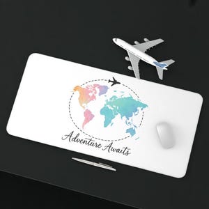 May include: White rectangular desk mat with a watercolor world map design in pastel colors. The text "Adventure Awaits" is below the map. A toy airplane and a computer mouse are on the mat.