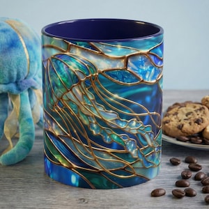 Puede incluir: Una taza de cerámica con un diseño de vidriera que presenta una medusa en tonos de azul, verde azulado y dorado. El interior de la taza es azul marino. Un peluche de medusa y un plato de galletas están en el fondo.