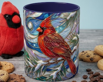 May include: A ceramic mug with a stained-glass design featuring a red cardinal perched on a snowy branch. The mug has a dark blue interior and is surrounded by cookies, coffee beans, and a plush cardinal toy.