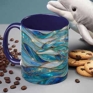 May include: A ceramic coffee mug with a blue interior and handle. The exterior features a stained-glass style design of a dolphin swimming in blue and teal waves. Coffee beans and cookies are in the background.