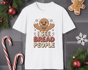 May include: White t-shirt with a cartoon gingerbread man design, a bow tie, and the text "I SEE BREAD PEOPLE". The design includes snowflakes and candy canes. A festive garment for the holiday season.