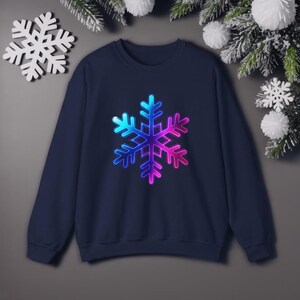 May include: Navy blue sweatshirt featuring a large, vibrant snowflake design. The snowflake transitions in color from blue to purple and pink. The sweatshirt is a crew neck style, perfect for winter or holiday wear.