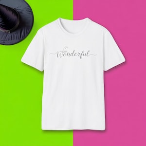Puede incluir: Camiseta blanca con la palabra "Wonderful" en cursiva, acompañada de un pequeño gráfico de sombrero de bruja. La camiseta está sobre un fondo verde y rosa vibrante, con un sombrero de bruja negro en la esquina superior izquierda.